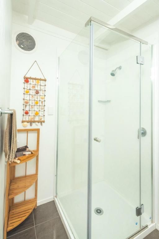 A glass-enclosed shower next to a wooden corner shelf holding toiletries, with a shell decoration hanging on the wall.