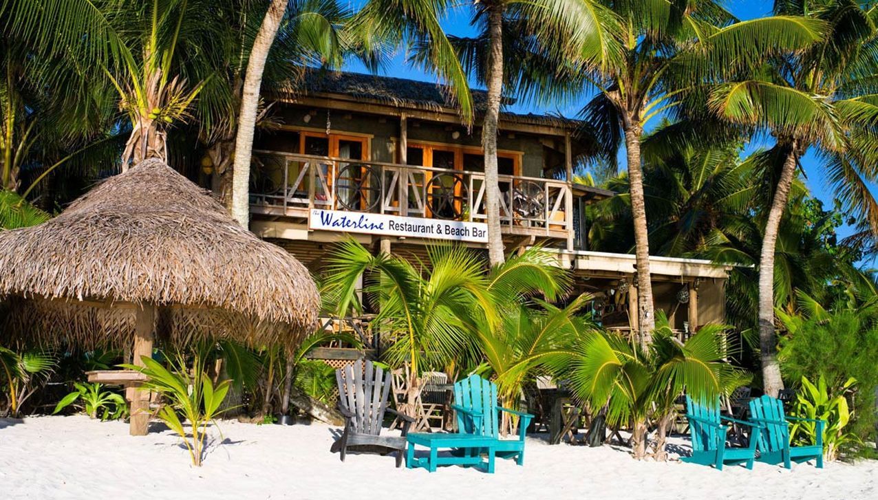 A two-story wooden building on a white sand beach with a thatched-roof tiki hut and turquoise chairs under palm trees.