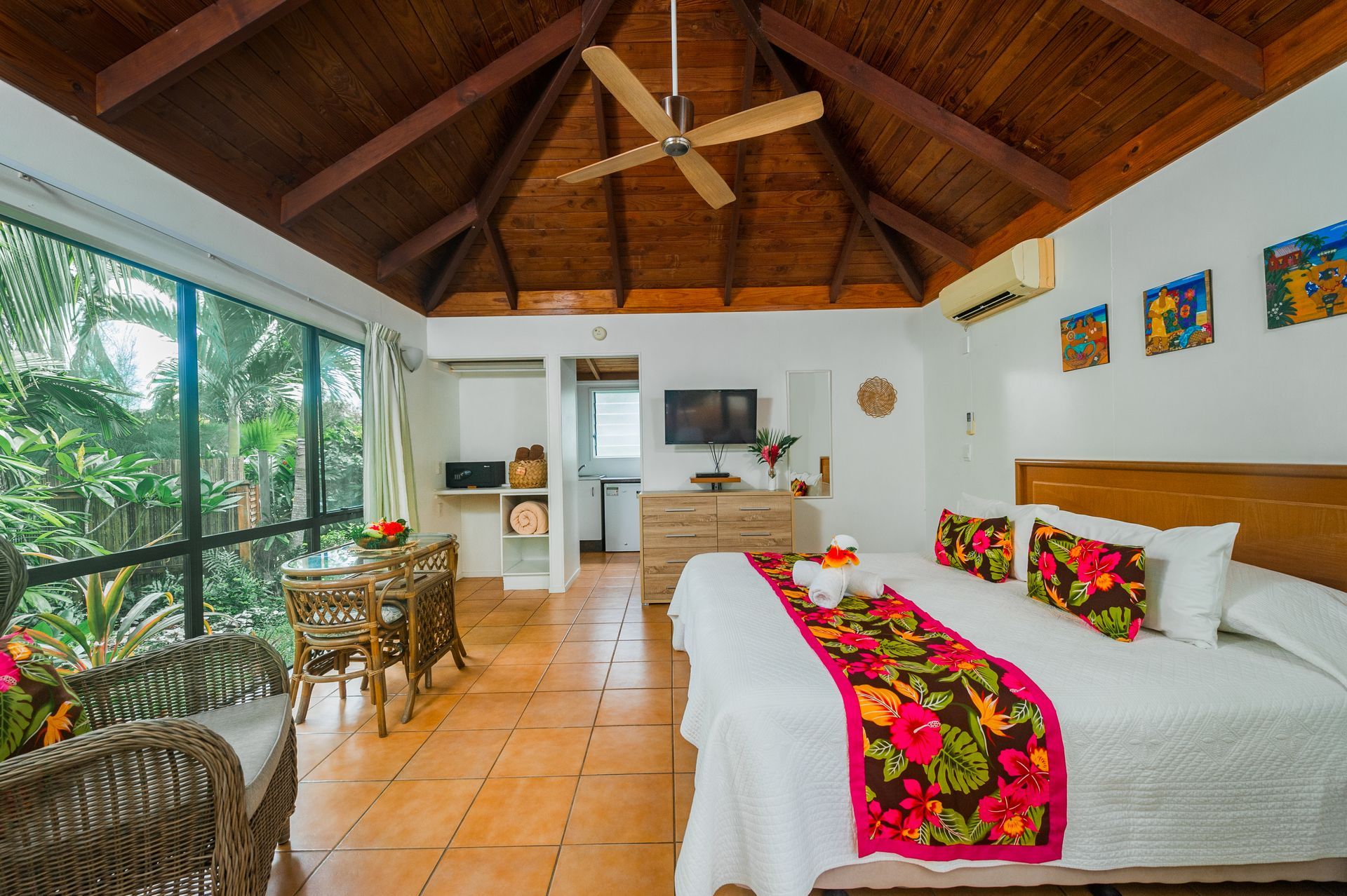 A tropical hotel room with a large bed, wooden vaulted ceiling, ceiling fan, wicker chairs, and floor-to-ceiling windows.