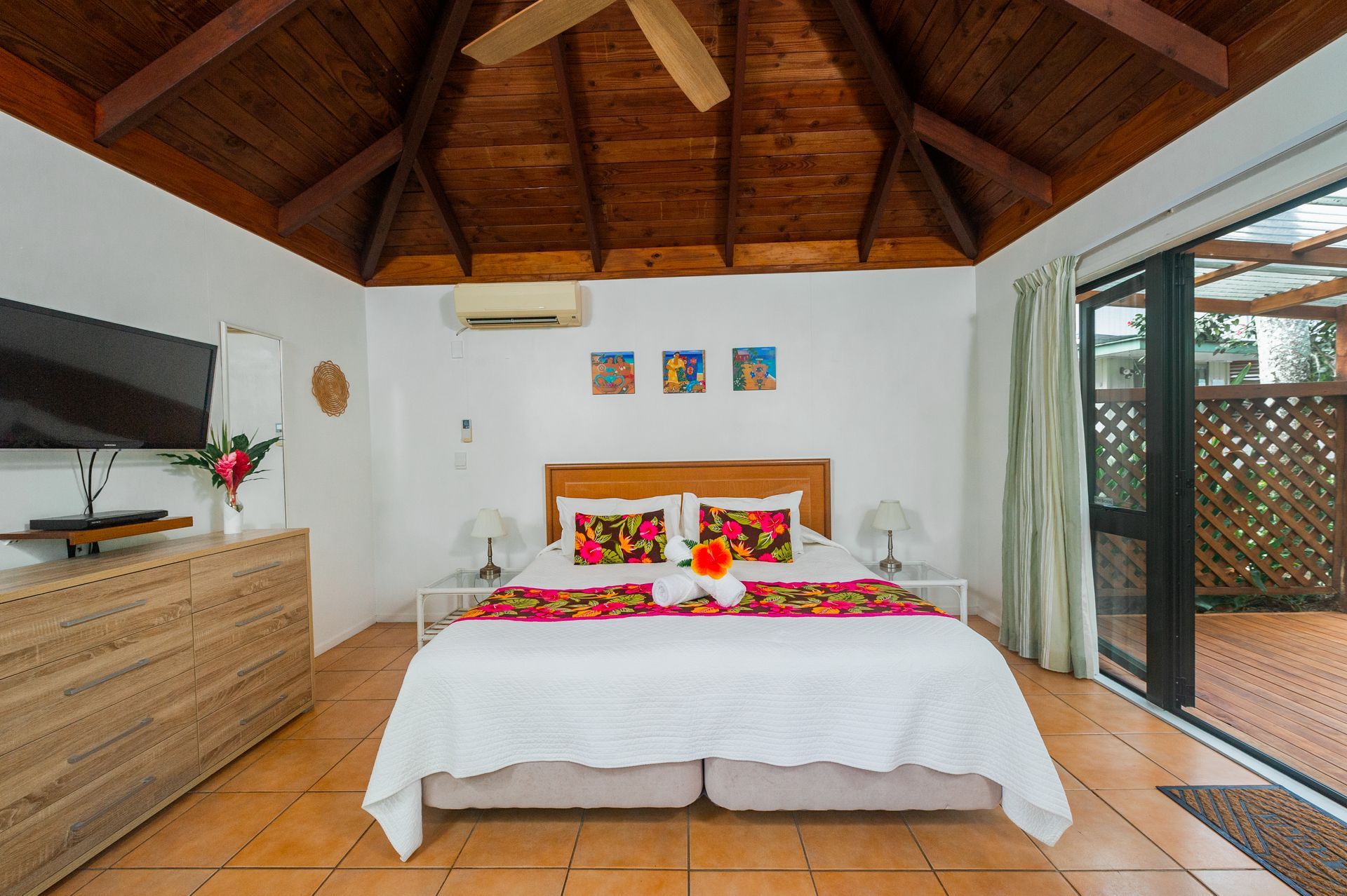 A bright bedroom with a king bed, wooden vaulted ceiling, tiled floors, a TV on a dresser, and a door to a patio.