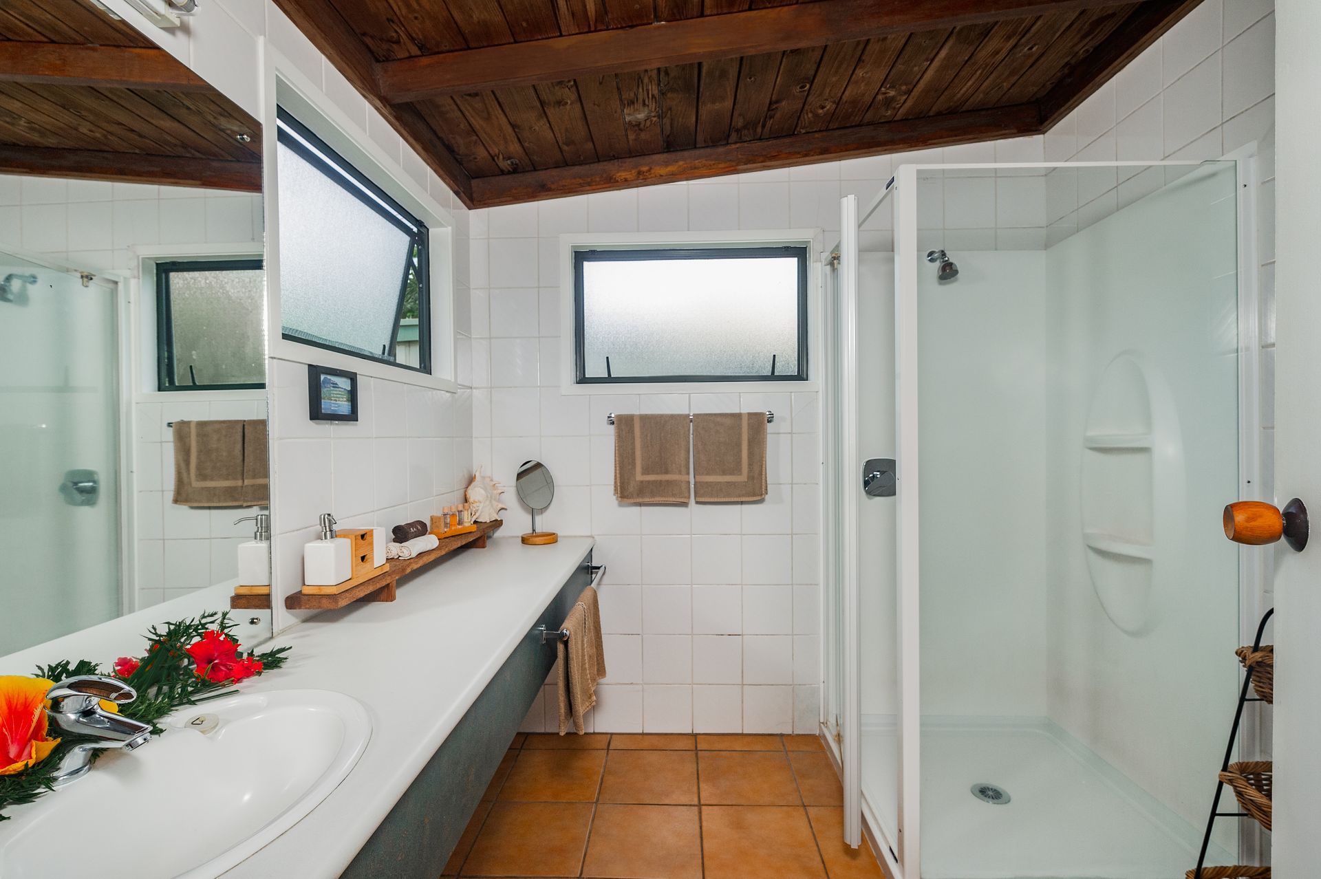 A bathroom with white tiled walls, a long vanity, a corner shower stall, and a dark wooden paneled ceiling.