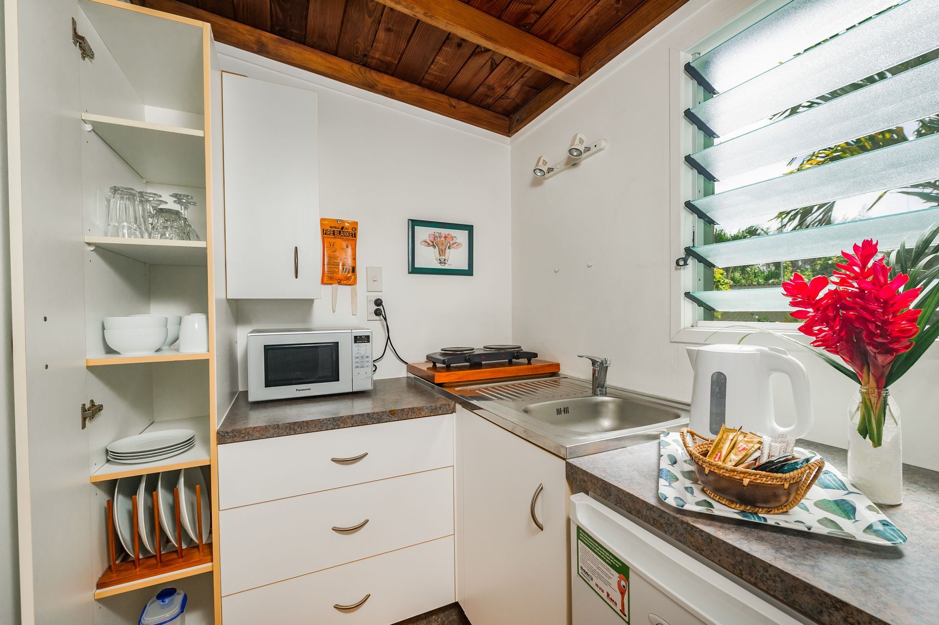 A compact kitchen with white cabinets, a microwave, sink, shelves with dishes, and a vase of bright red flowers.