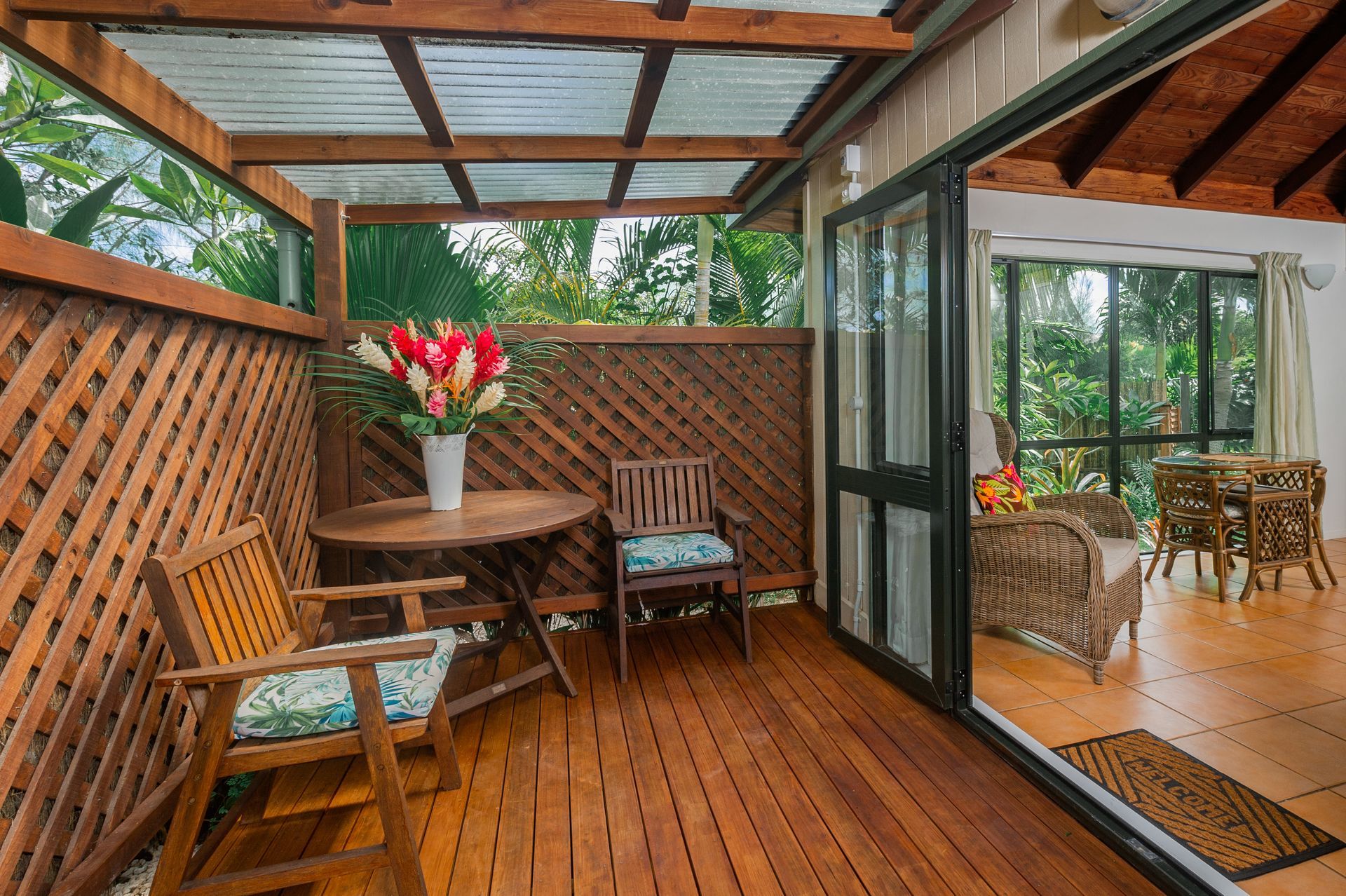 A covered wooden deck features a round table, two chairs, and a sliding door leading to an interior living space.