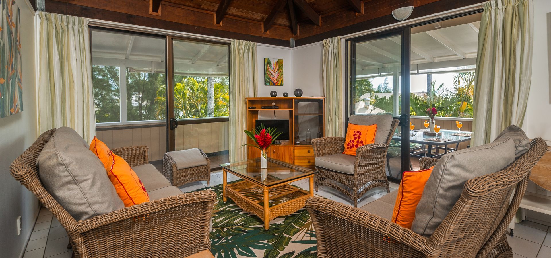 A sunroom with wicker furniture, orange accent pillows, a wooden coffee table, and large windows looking onto greenery.
