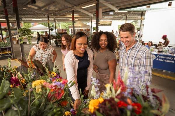 Three people smiling and looking at flowers at an outdoor market stall
