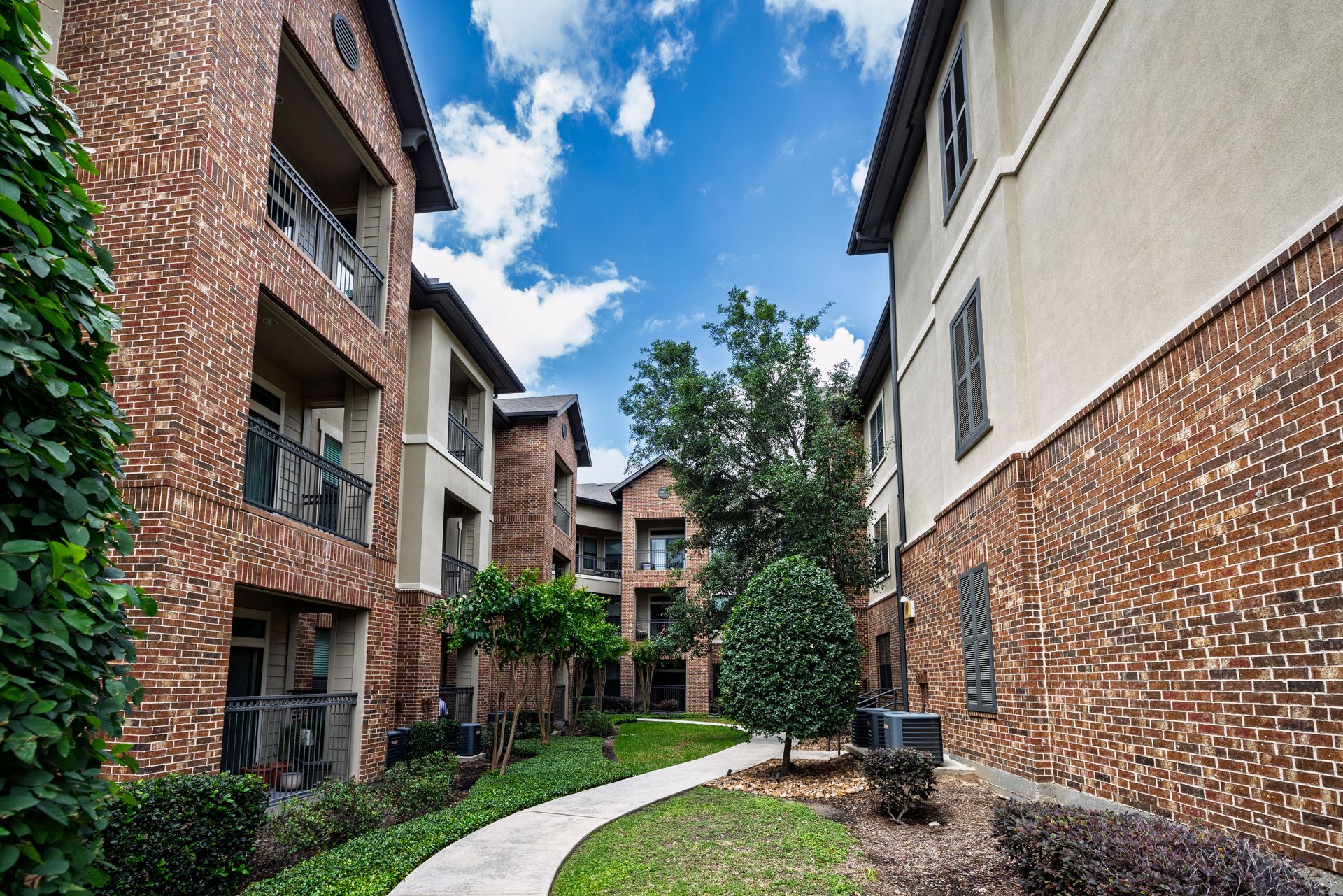 Courtyard path between brick apartment buildings under a blue sky at Olympus Sierra Pines at The Woodlands, TX
