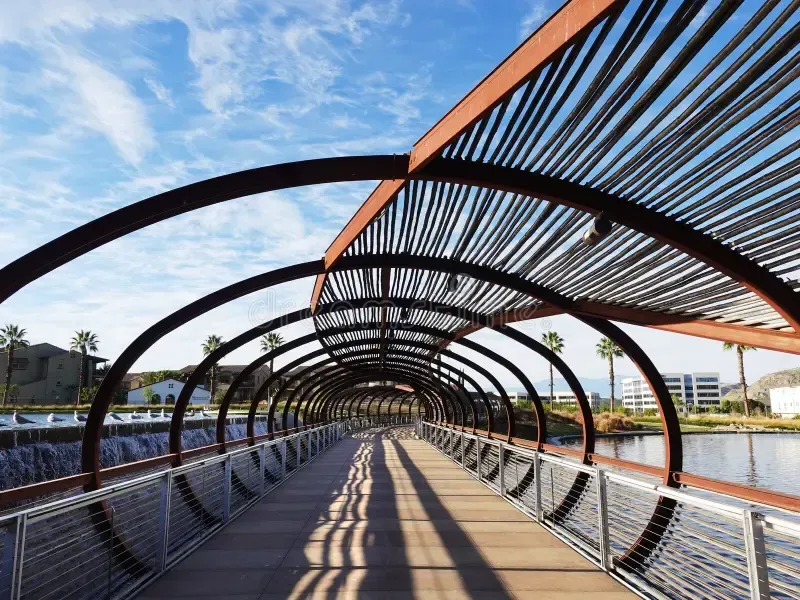 Modern pedestrian bridge with arched black beams and orange accents over water, casting long shadows under a blue sky
