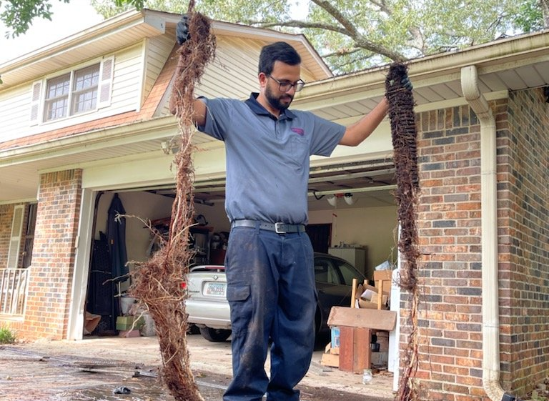 Faucet Installation Man holding two long root masses from a house's gutter. He stands in front of the house and garage.