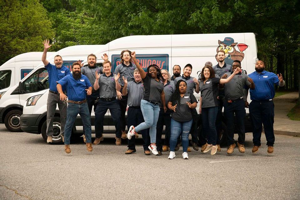 Group of people jumping in front of company vans, smiling, waving. Outdoors, day.