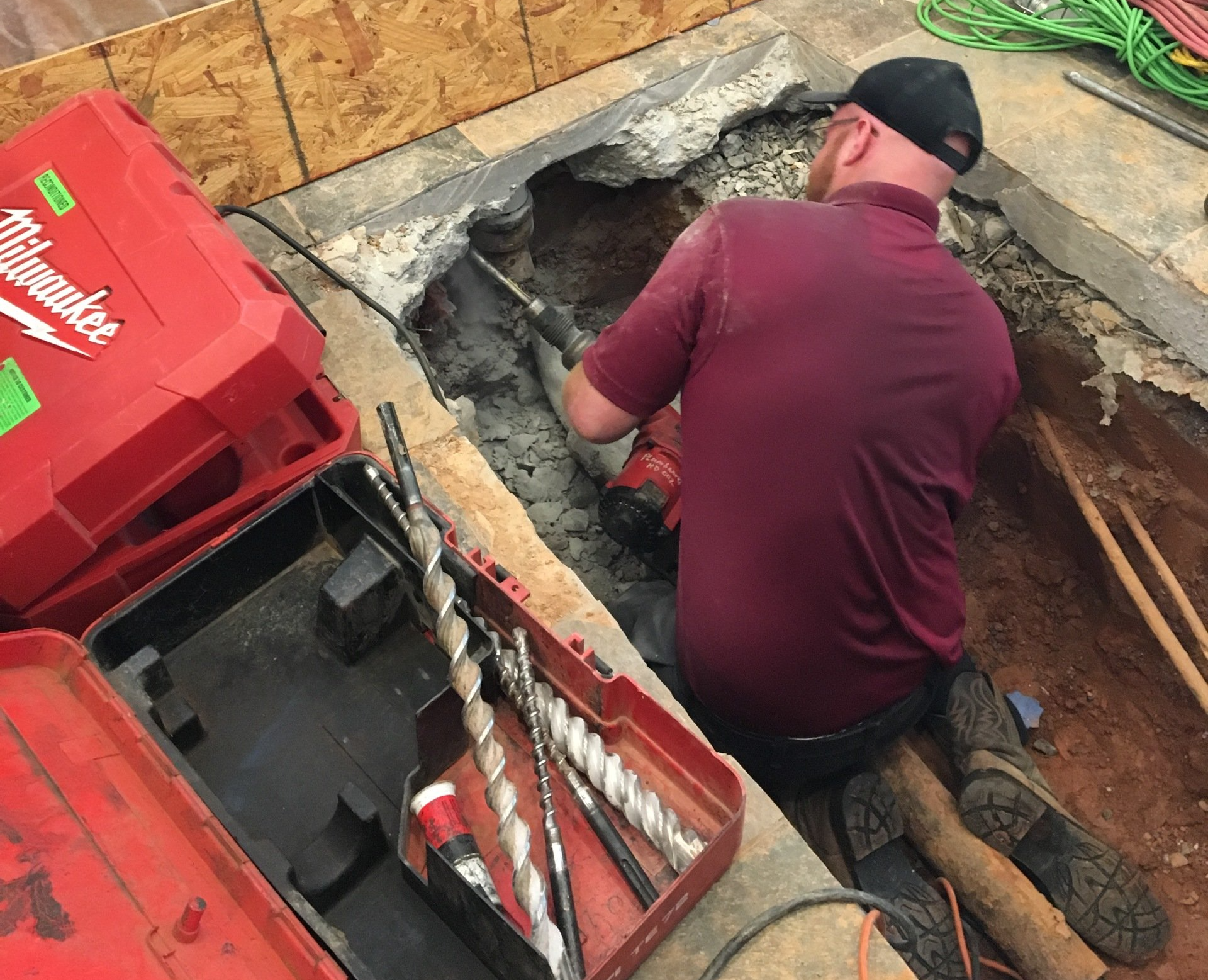 Man using a power drill in a concrete hole with a toolbox and drill bits nearby.