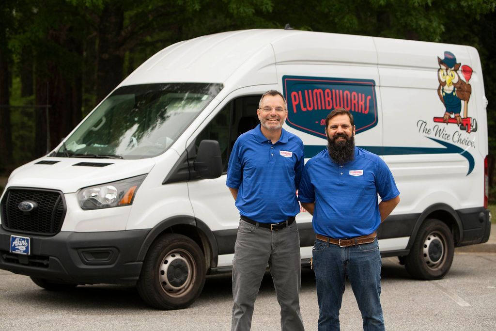 Two men in blue shirts stand beside a white plumbing van with logo.