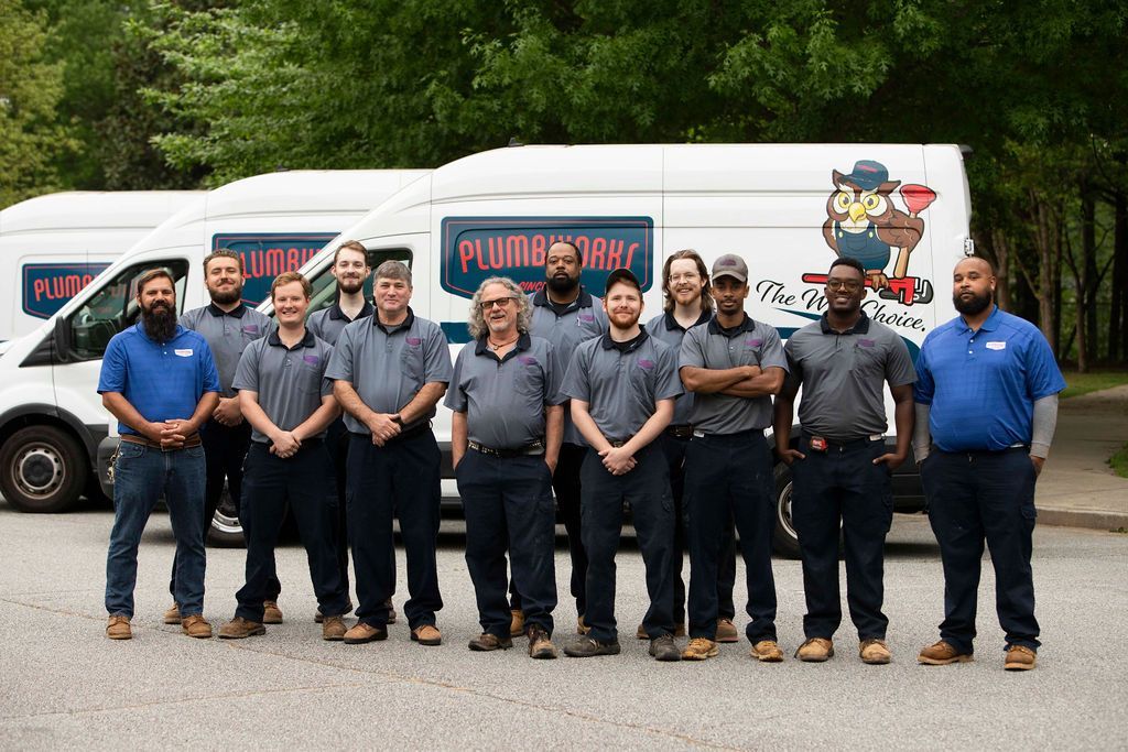 Plumbers standing in front of van with logo. They wear matching blue and gray shirts and pants, posing outside.