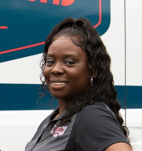 Woman with wavy hair, smiling, wearing a work shirt, standing in front of a blue and white vehicle.