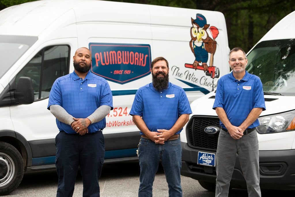 Three plumbers in blue shirts pose in front of white vans with company logo.