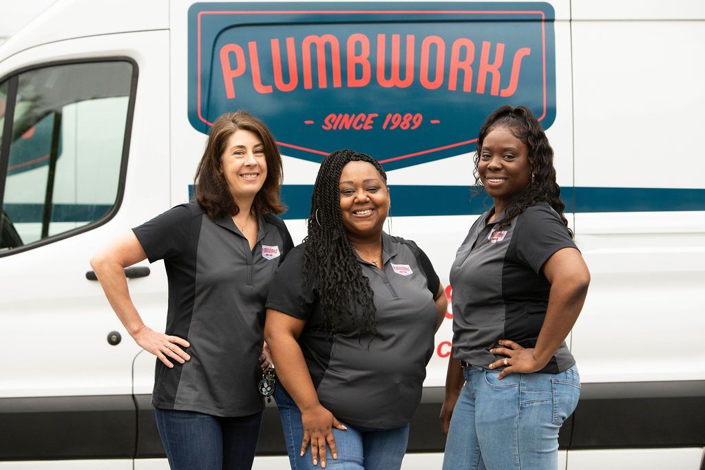 Three women in black shirts in front of a white Plumbworks truck; they are smiling.