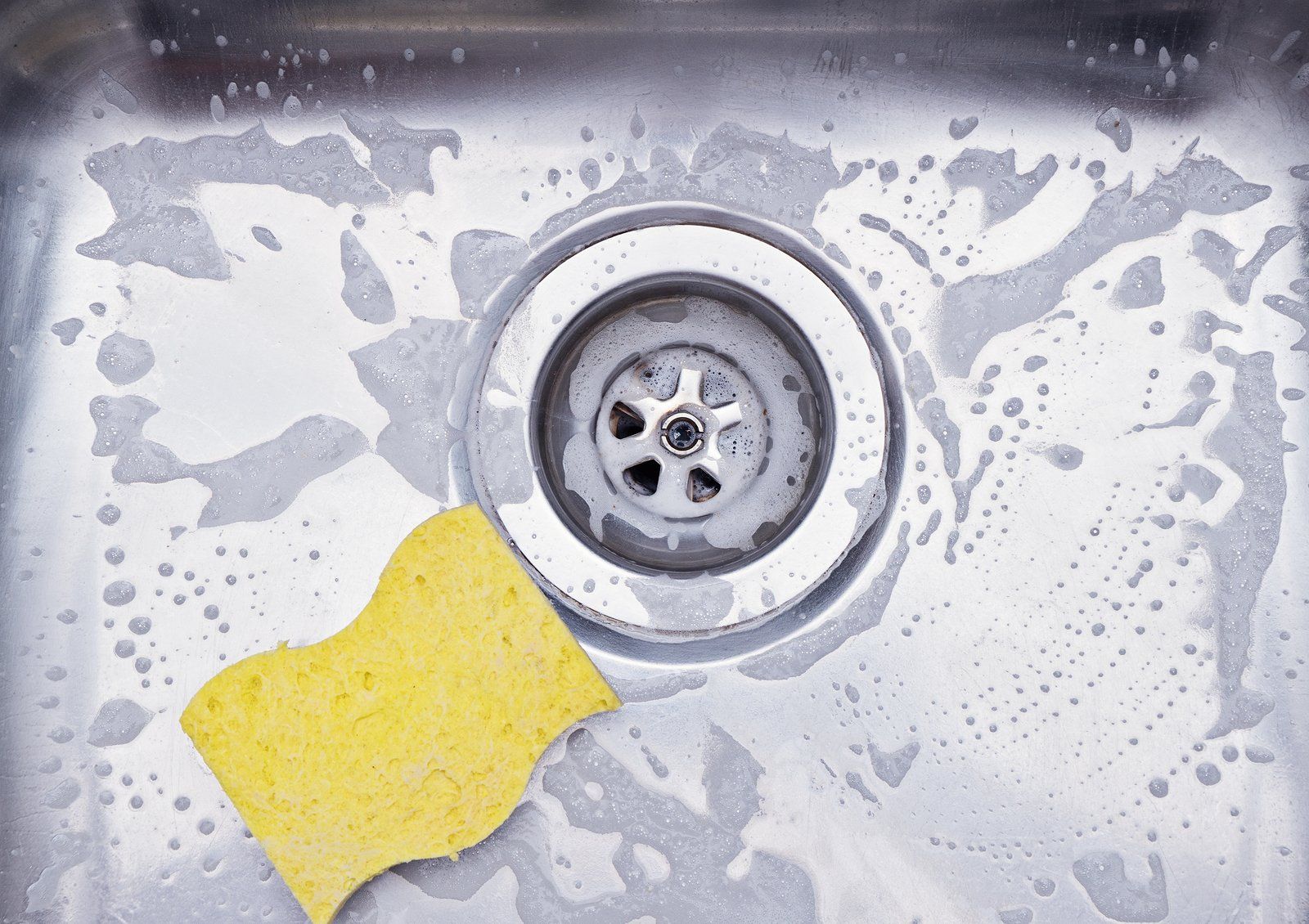 Drain Cleaning Yellow sponge cleaning a stainless steel kitchen sink with soap suds around the drain.