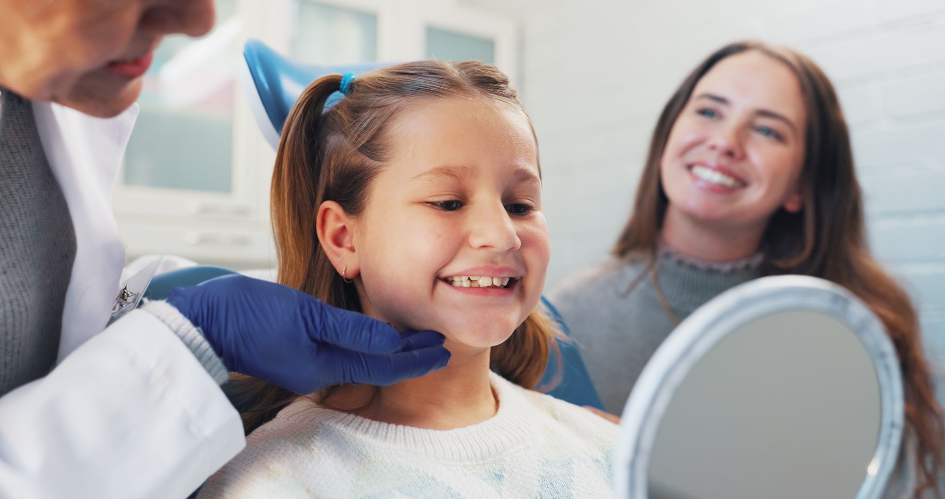 Dentist examining a smiling child in a clinic, with a mother watching in the background.
