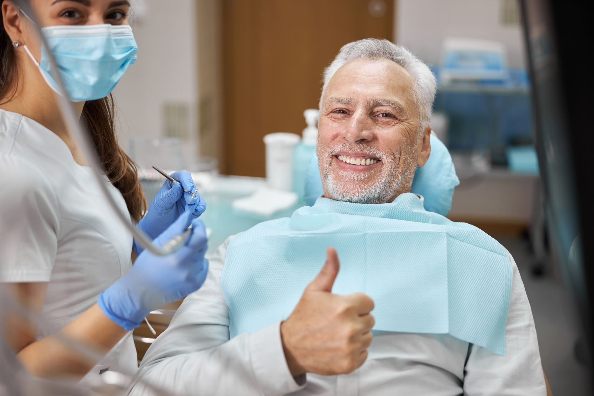 Dentist and masked assistant in a clinic; smiling patient gives a thumbs-up in the chair.