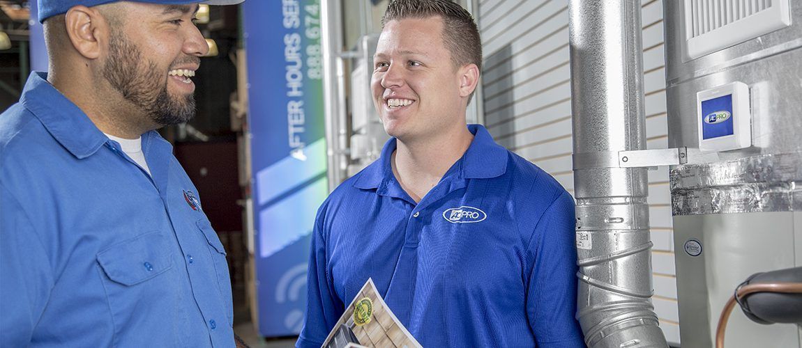Two men in blue work shirts talking in an industrial hallway, one holding a brochure.