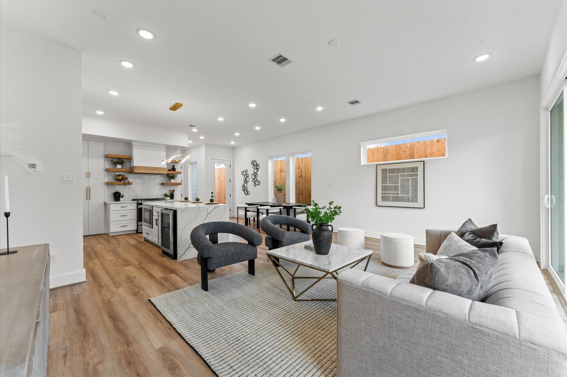 Modern, bright open-concept living room with kitchen and seating area; light wood floors, white walls, gray furniture.