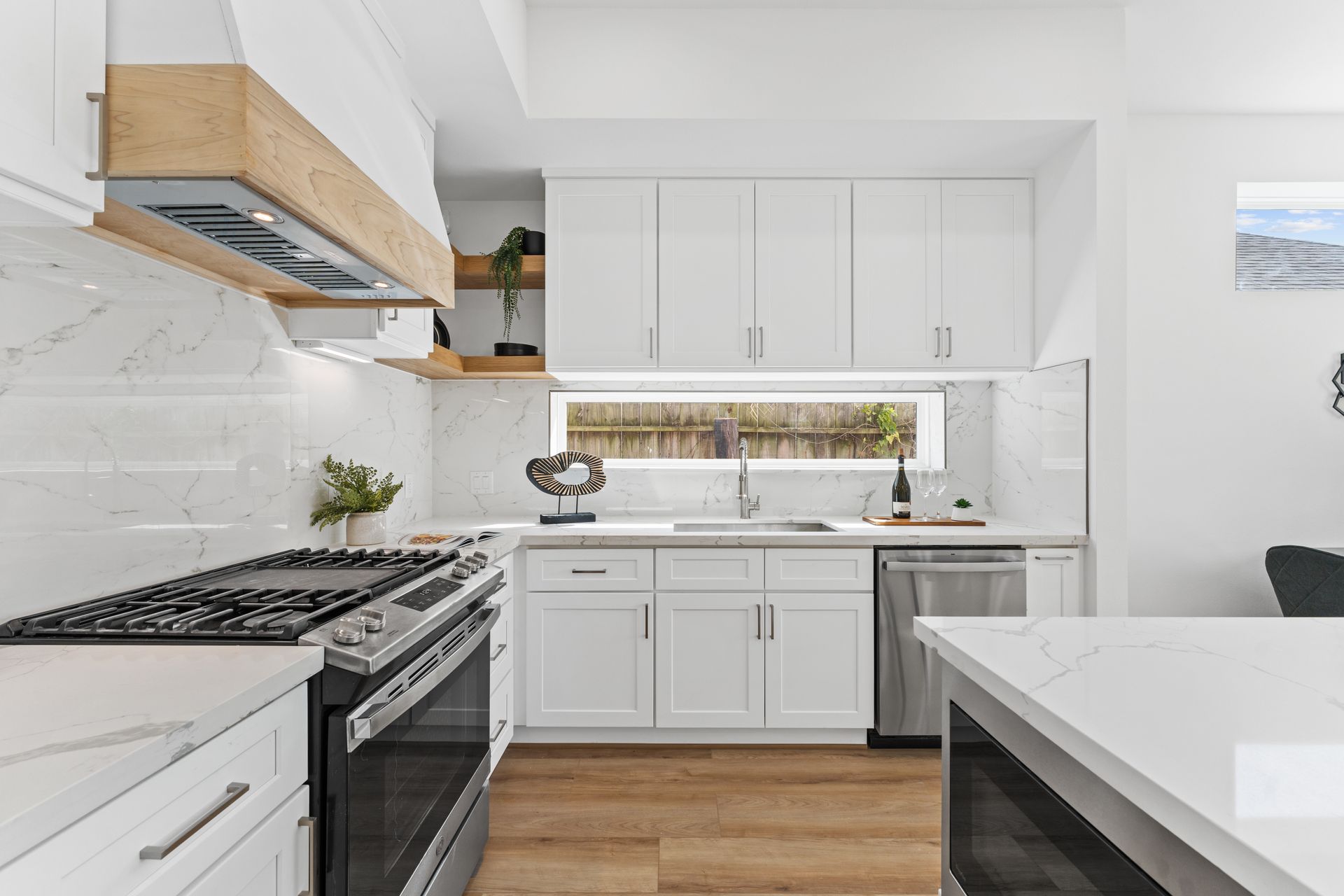 Bright white kitchen with marble countertops, stainless steel appliances, and a wooden range hood.