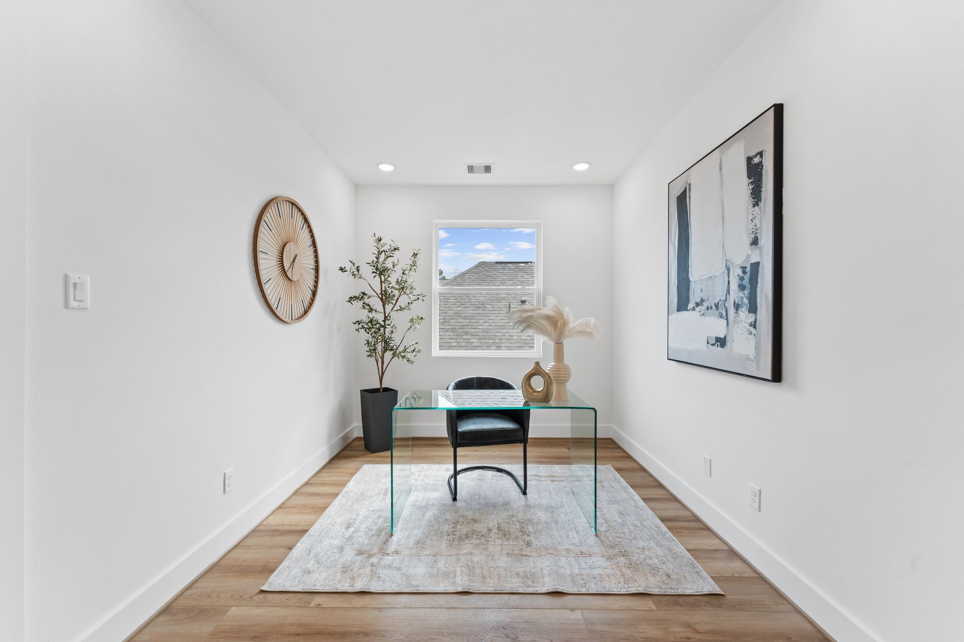 A home office with a glass desk, art, and a window overlooking a landscape. White walls and hardwood floors.