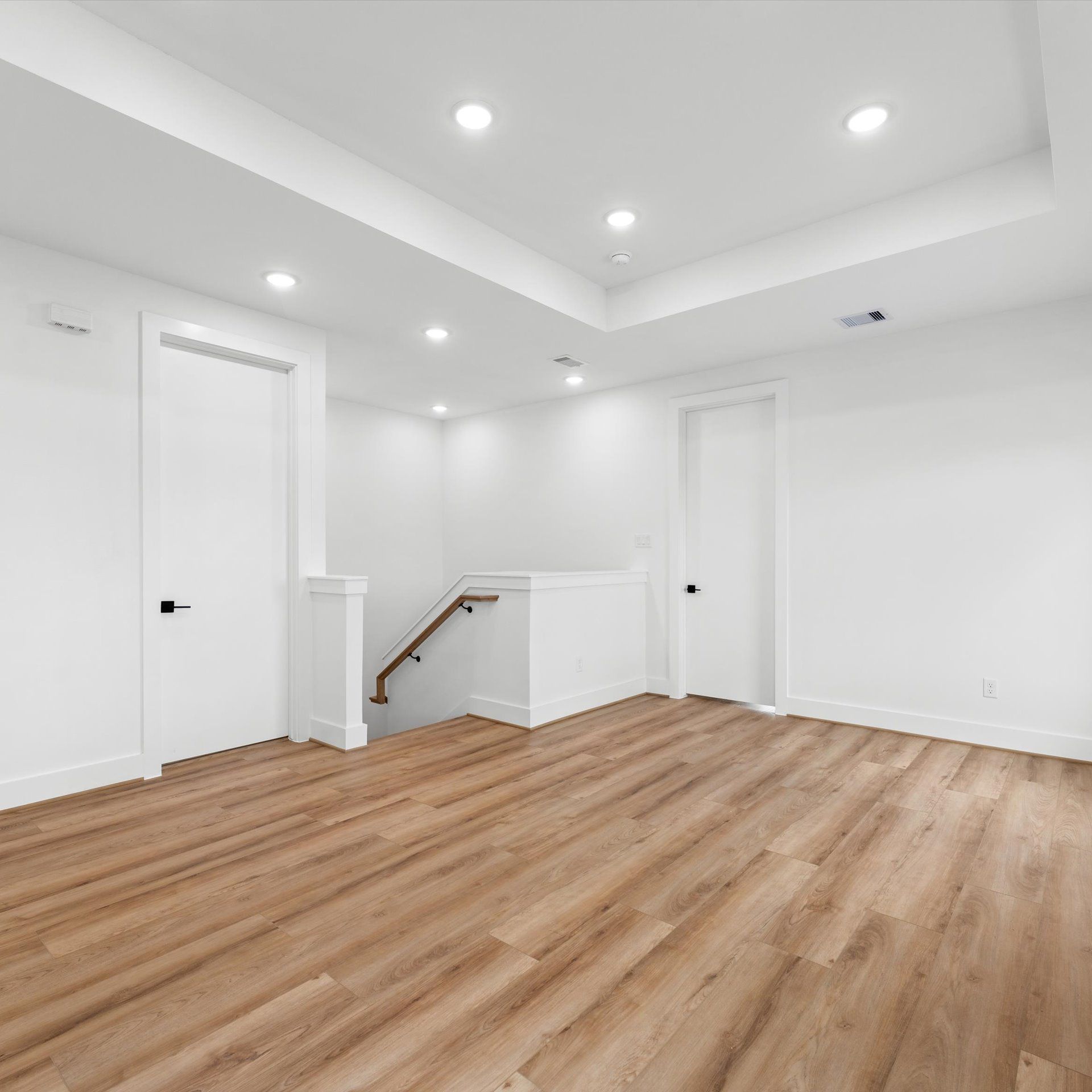 Empty room with wooden floors, white walls, recessed lighting, and a staircase.