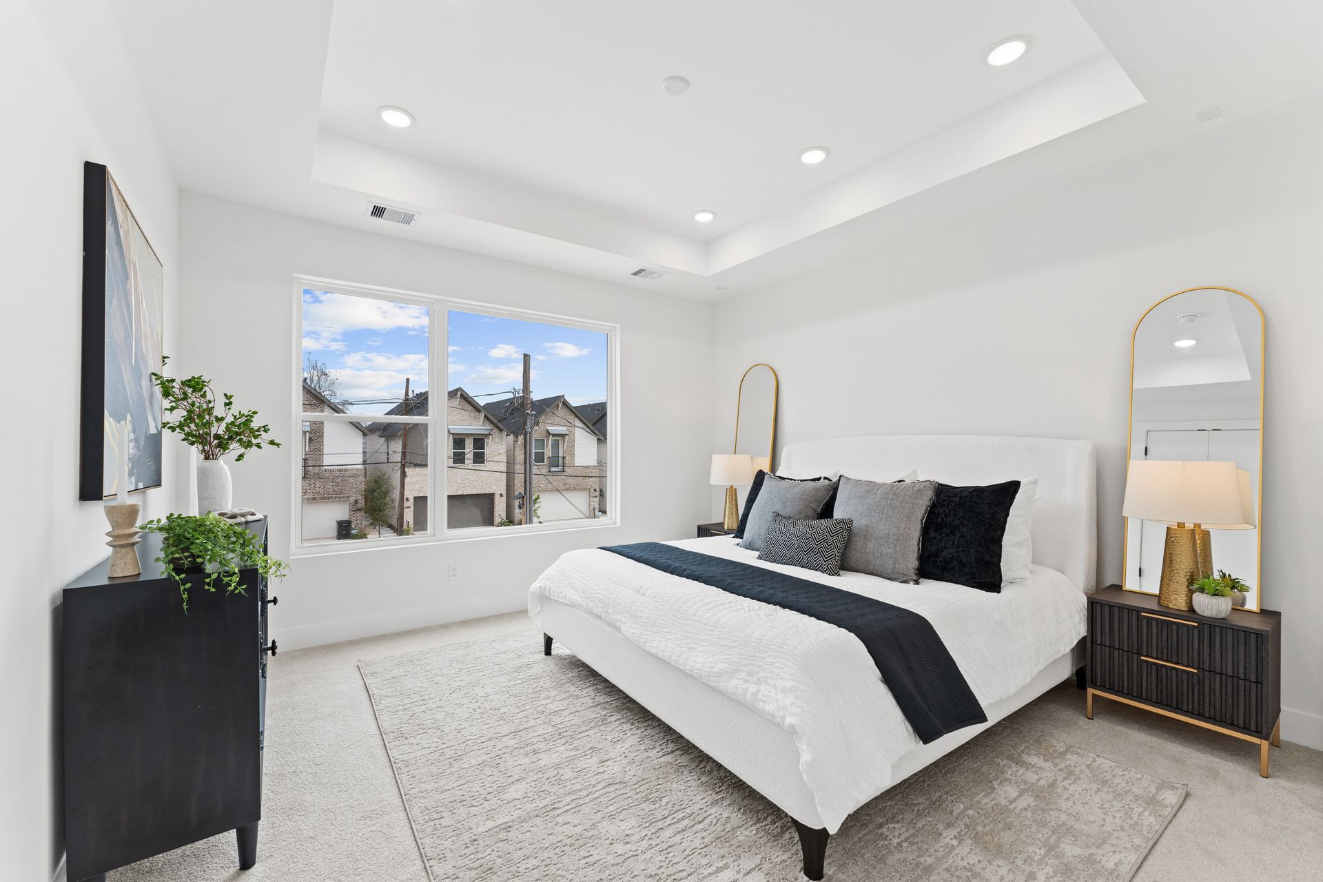 Bright bedroom with a white bed, black dresser, and large window overlooking a neighborhood.