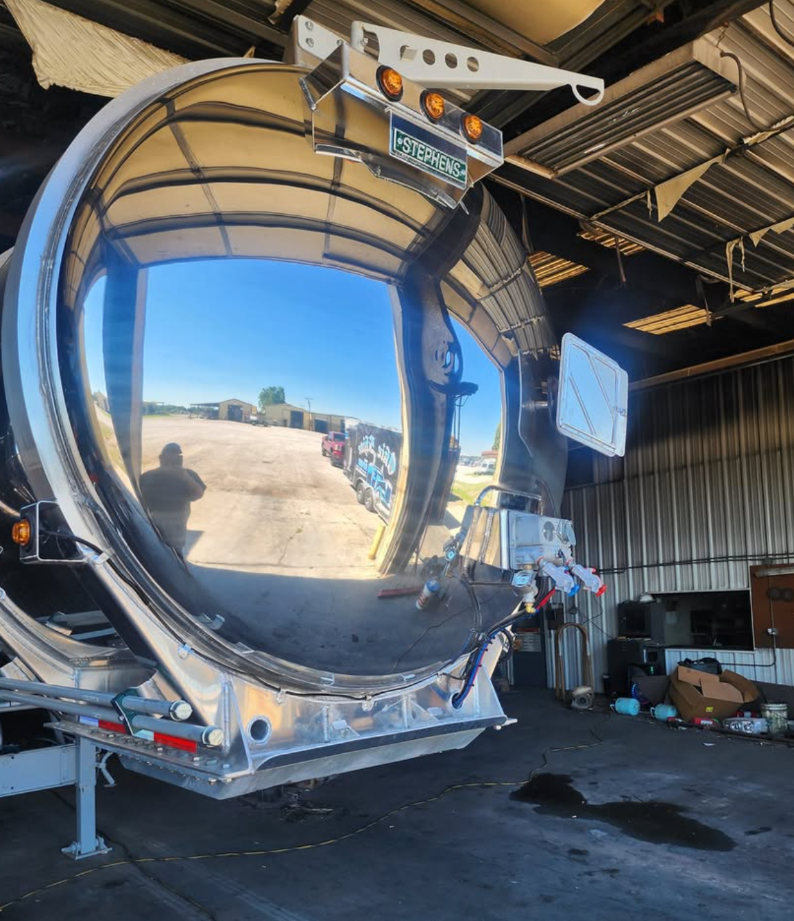 Shiny, reflective tanker truck with blue sky reflected in its surface. A person is visible in the reflection.