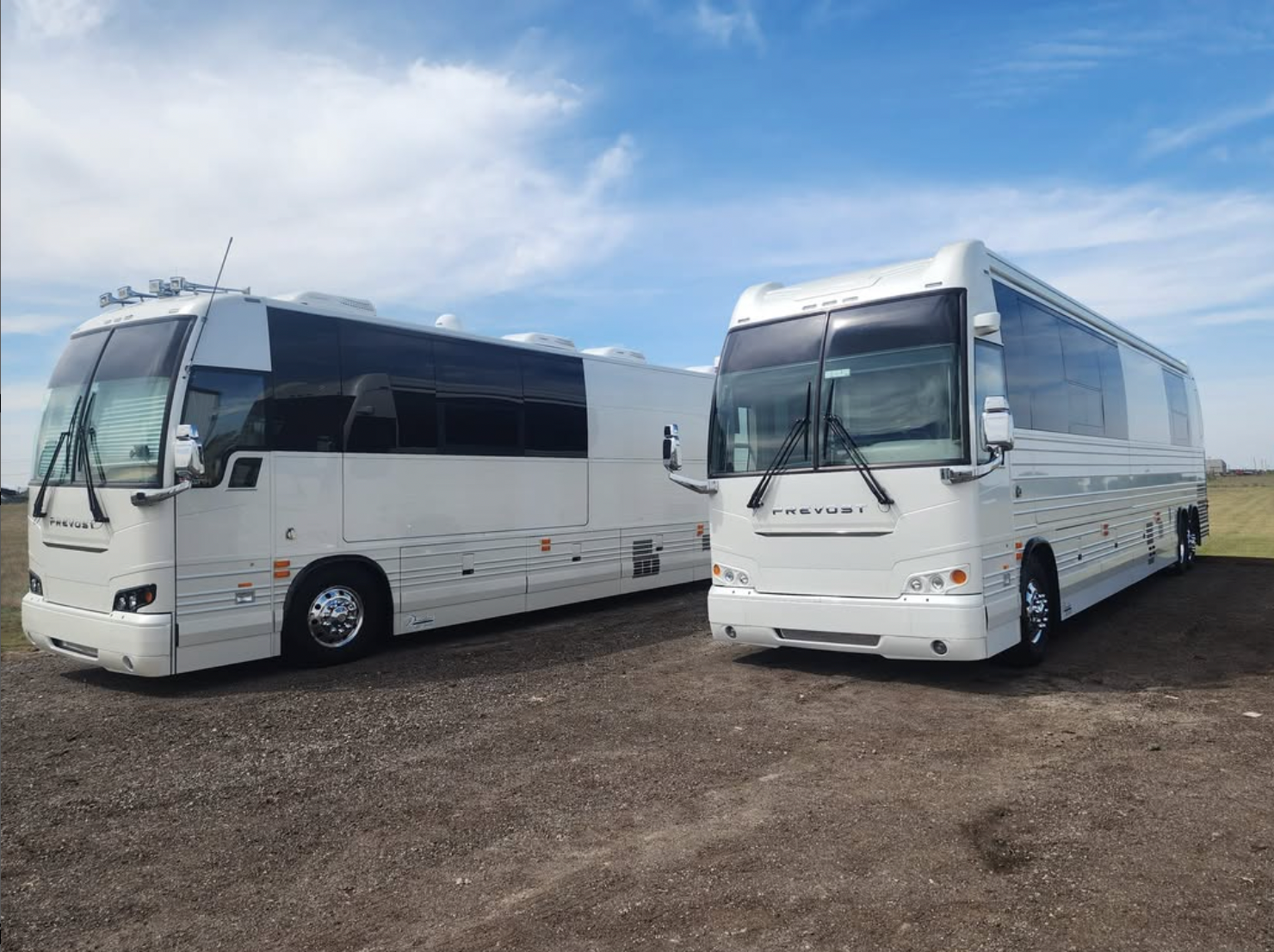 Two white tour buses parked outdoors on a gravel lot under a blue sky.