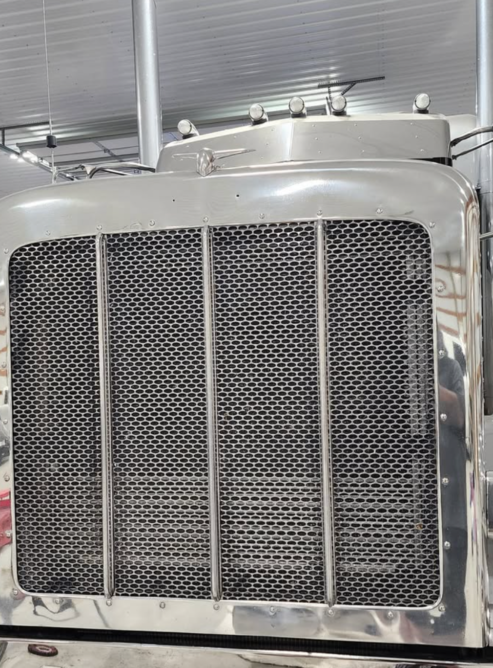 Chrome grill and hood of a semi-truck, with a mesh insert, under a metal ceiling.