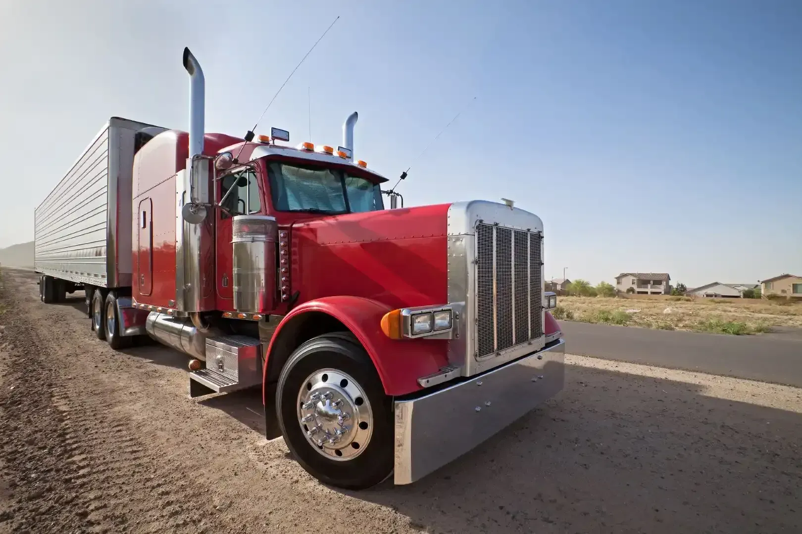 Red semi-truck parked on a dirt road next to pavement, under a blue sky.