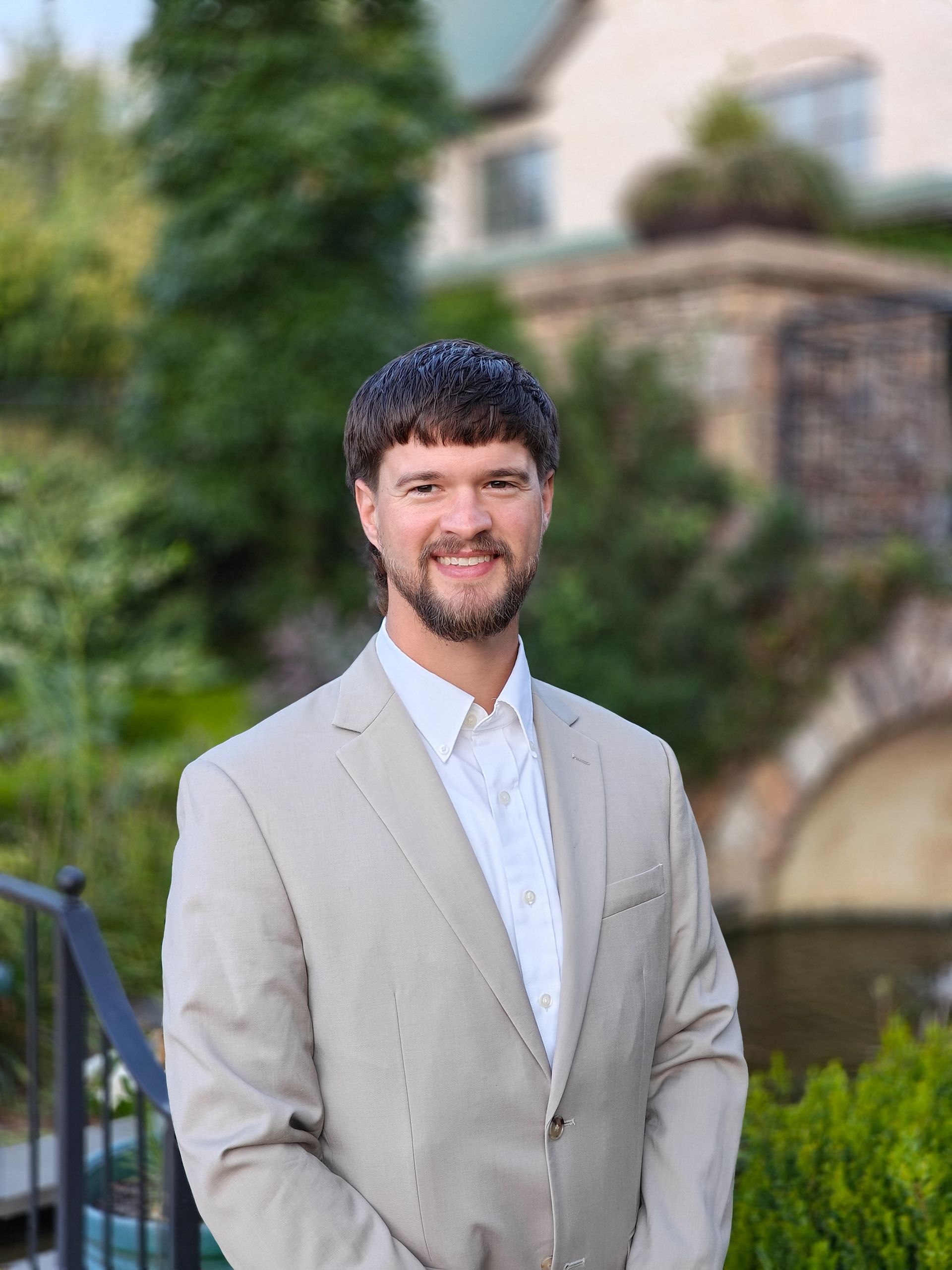 Man in a light beige suit smiling outdoors in front of a stone building and greenery