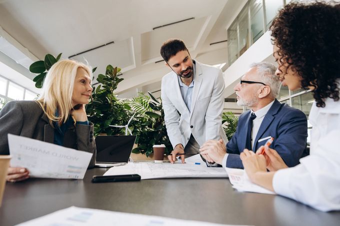 Team meeting around a table in a bright office, with one person presenting documents to colleagues.