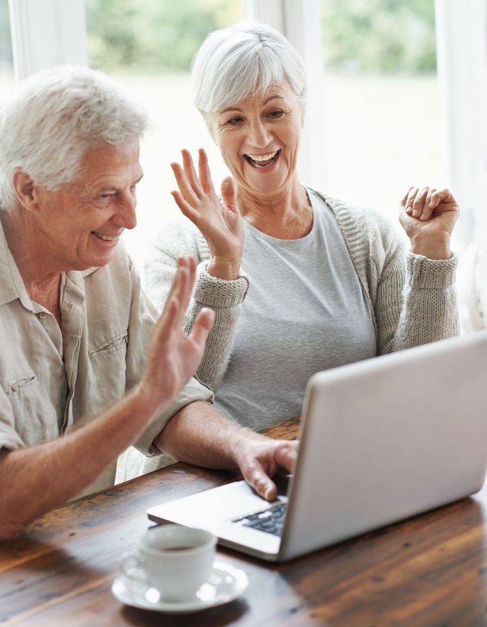 Two people smiling and waving at a laptop during a video call at a wooden table