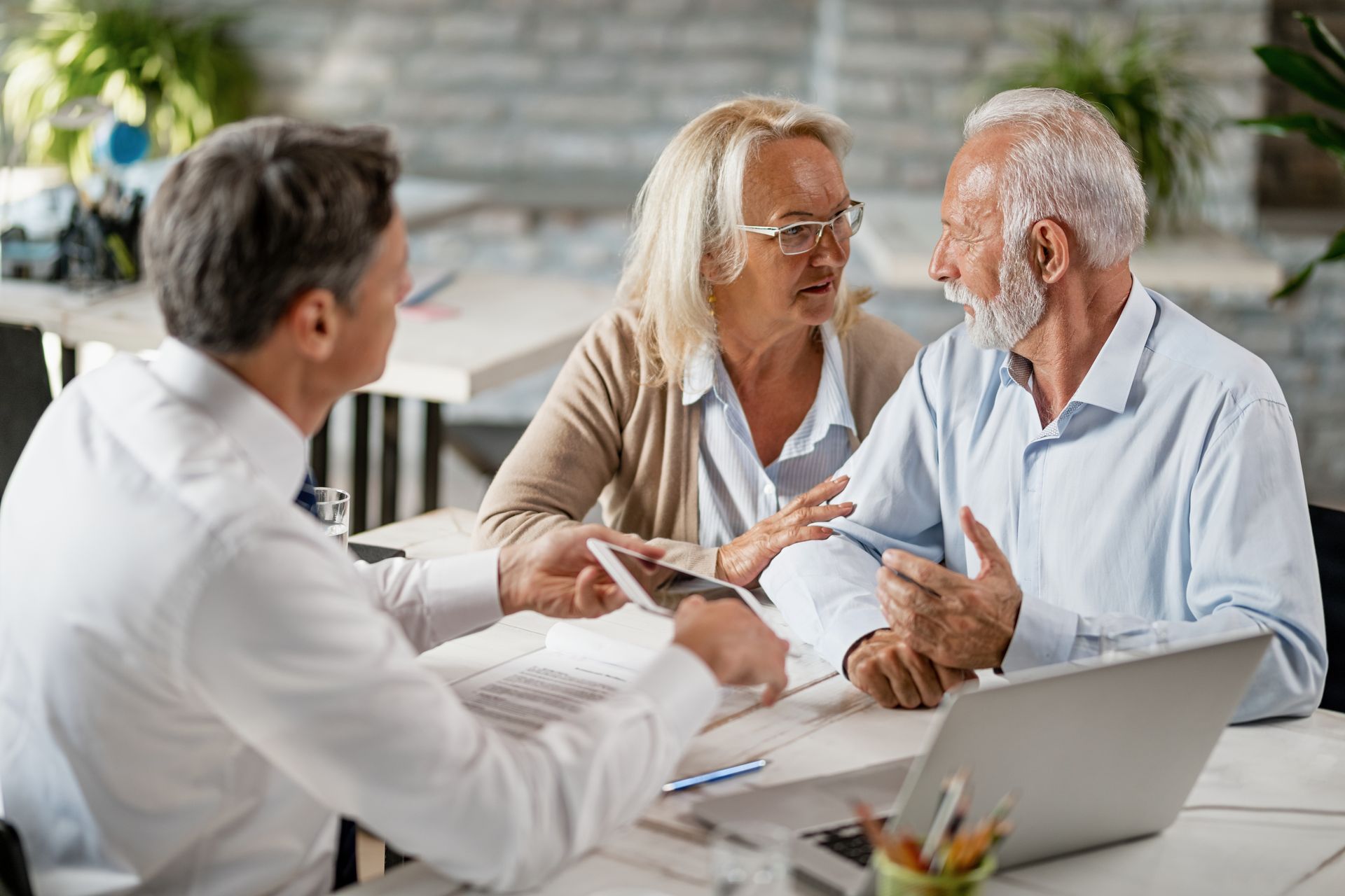 Three people in a bright office meeting, talking over papers beside a laptop.
