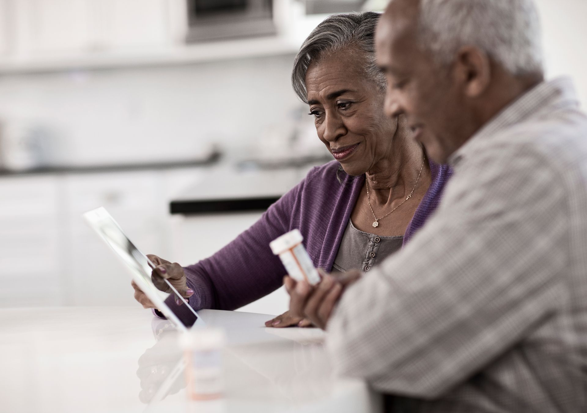Two older adults smiling at a tablet and medicine bottle on a kitchen table