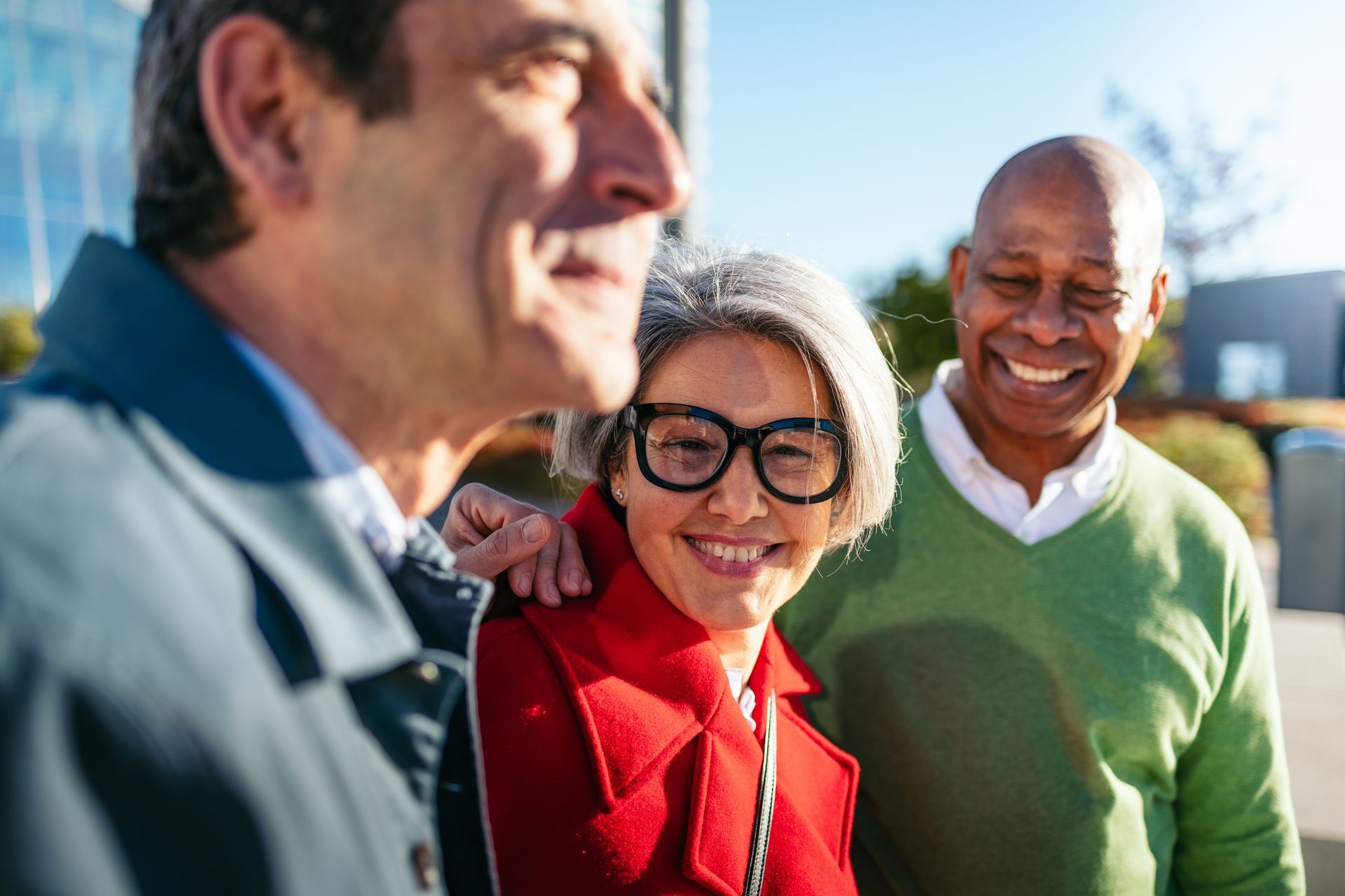 Three people smiling outdoors in bright sunlight, with one person in a red jacket in focus.
