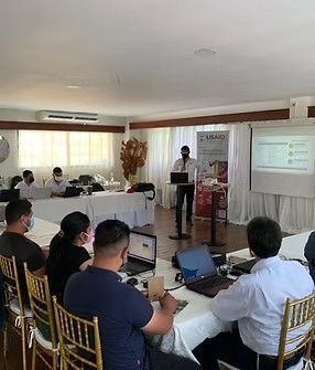 Un hombre realiza una presentación en una capacitación, los participantes se sientan en mesas con computadoras portátiles.