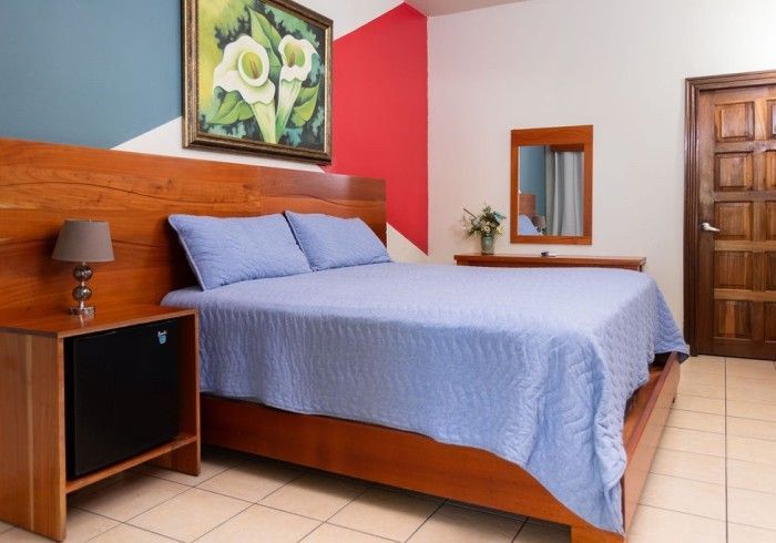 Dormitorio con cama de madera, ropa de cama azul, obras de arte, mesita de noche y puerta. Paredes azules, blancas y rojas.
