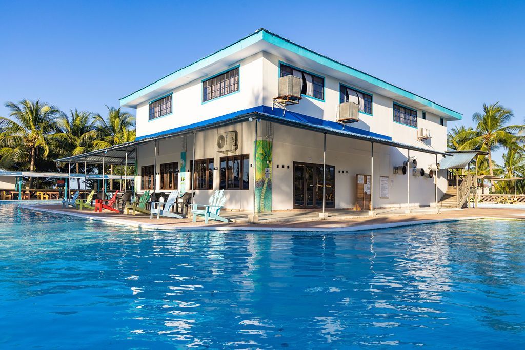 Un edificio blanco de dos pisos con detalles azules se encuentra junto a una gran piscina azul bajo un cielo despejado.