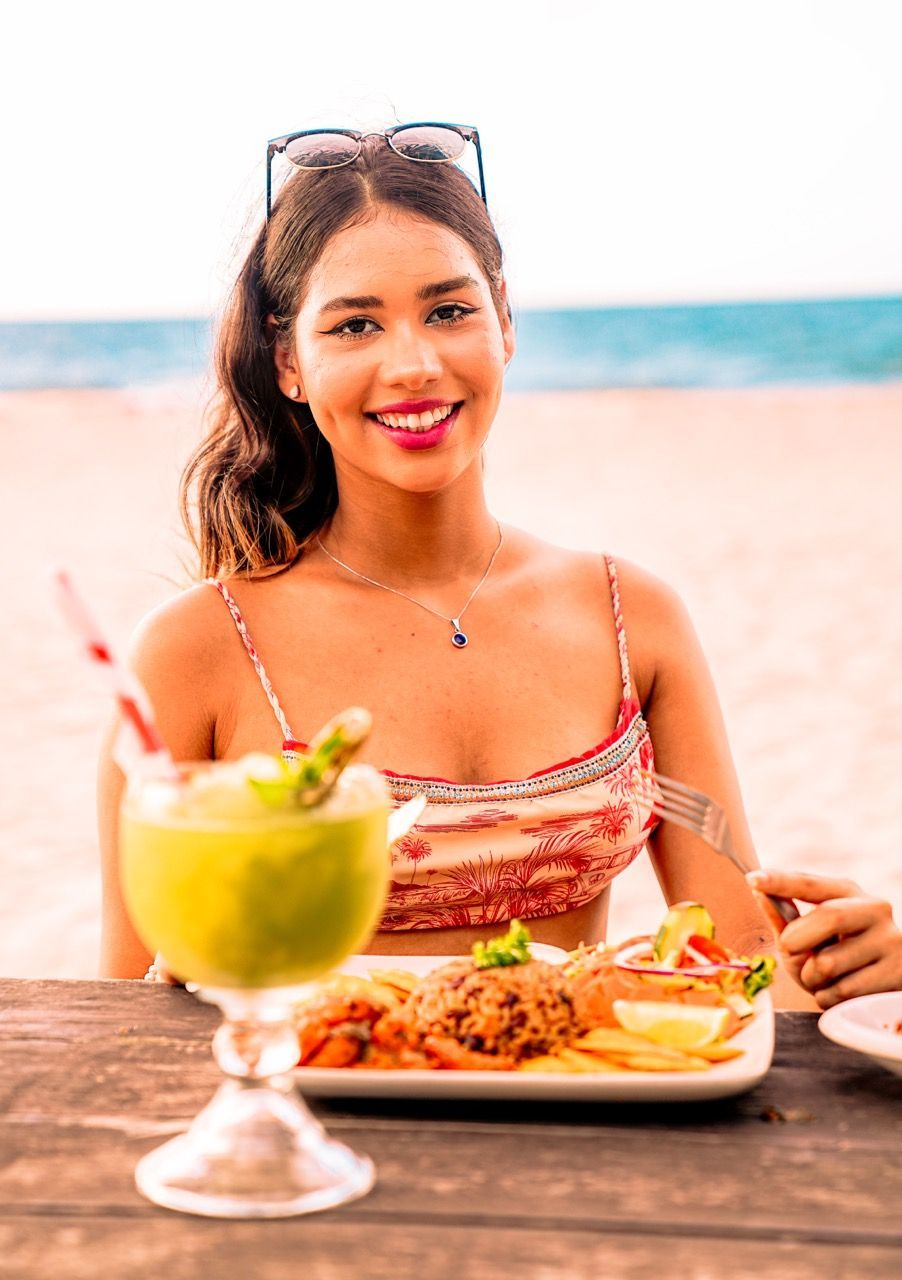 Mujer en una mesa junto a la playa con comida y bebida; sonriendo.