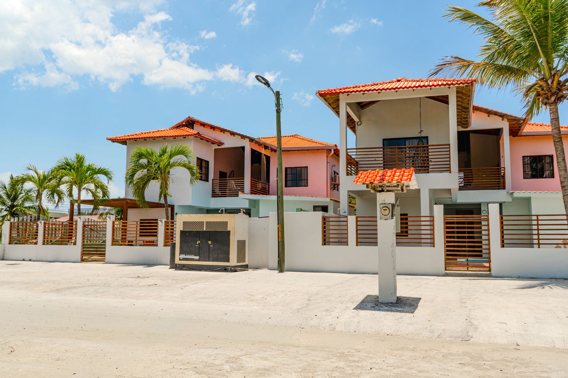 Casas de dos pisos con paredes rosas y blancas, techos de tejas naranjas, palmeras y una calle.