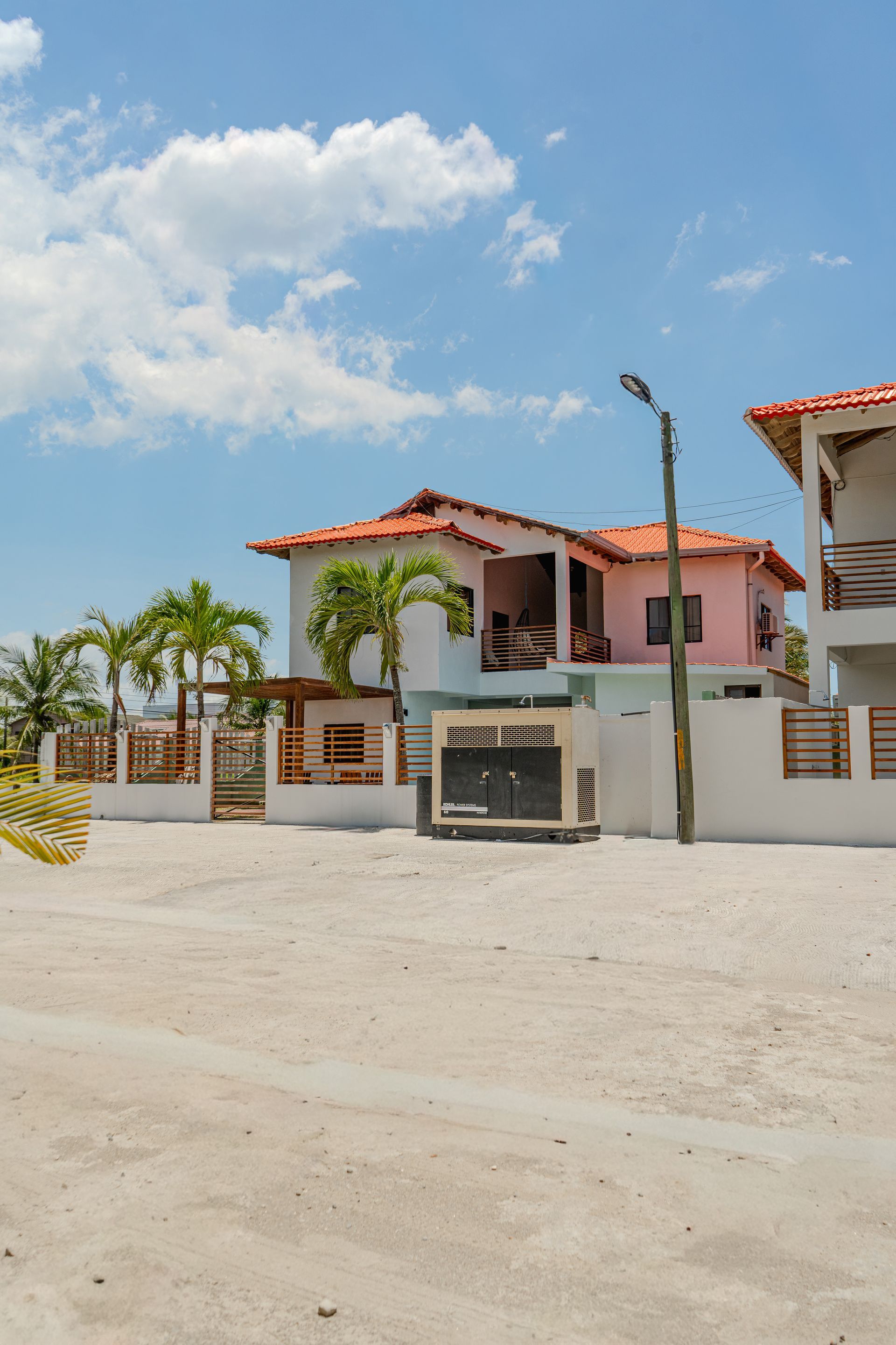 Casas de playa rosas y blancas con techos naranjas bajo un cielo azul brillante. Palmeras y suelo arenoso.