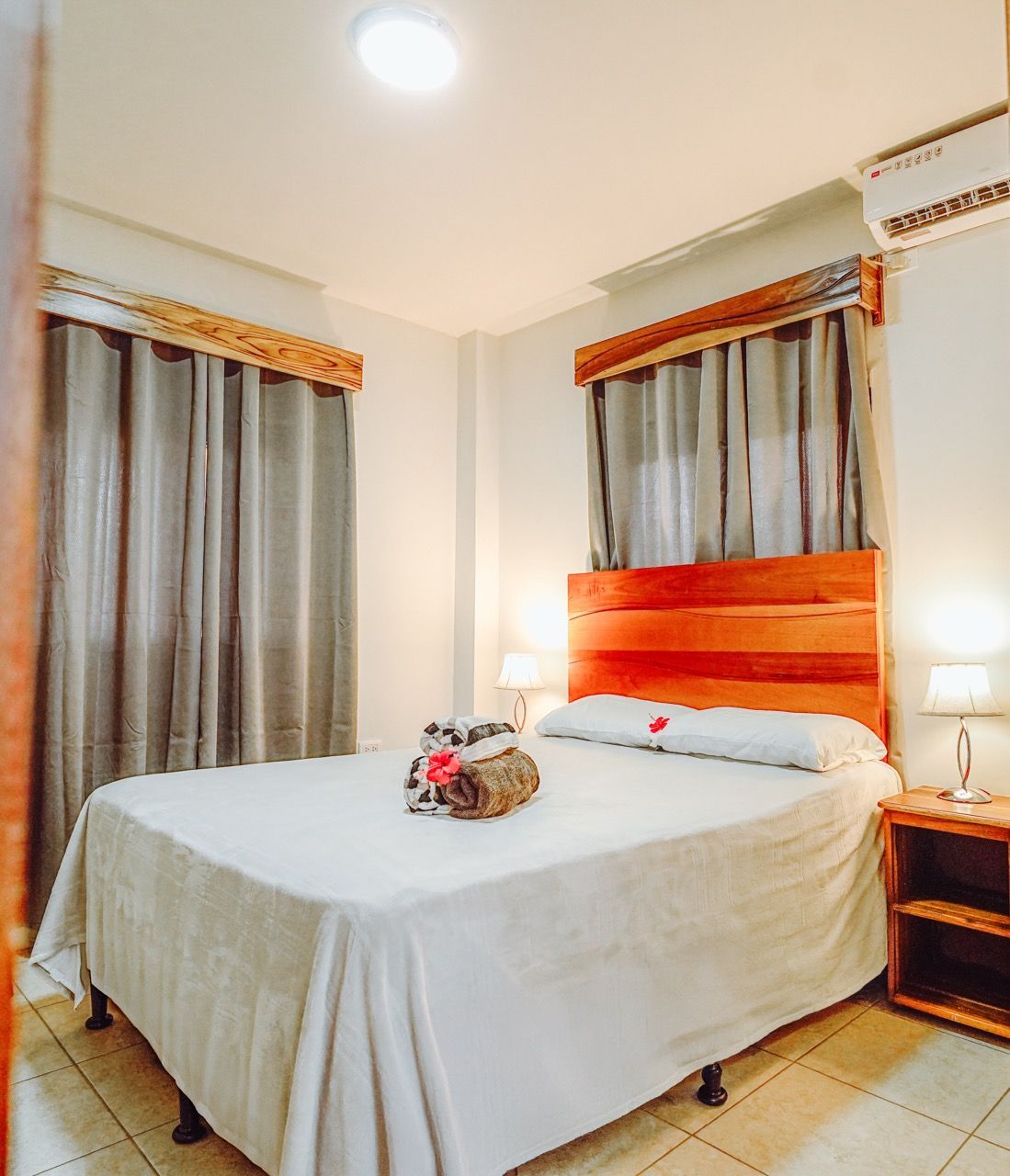 Dormitorio con cama, mesita de noche y cortinas. Las paredes son blancas, con cabecero de madera y molduras.