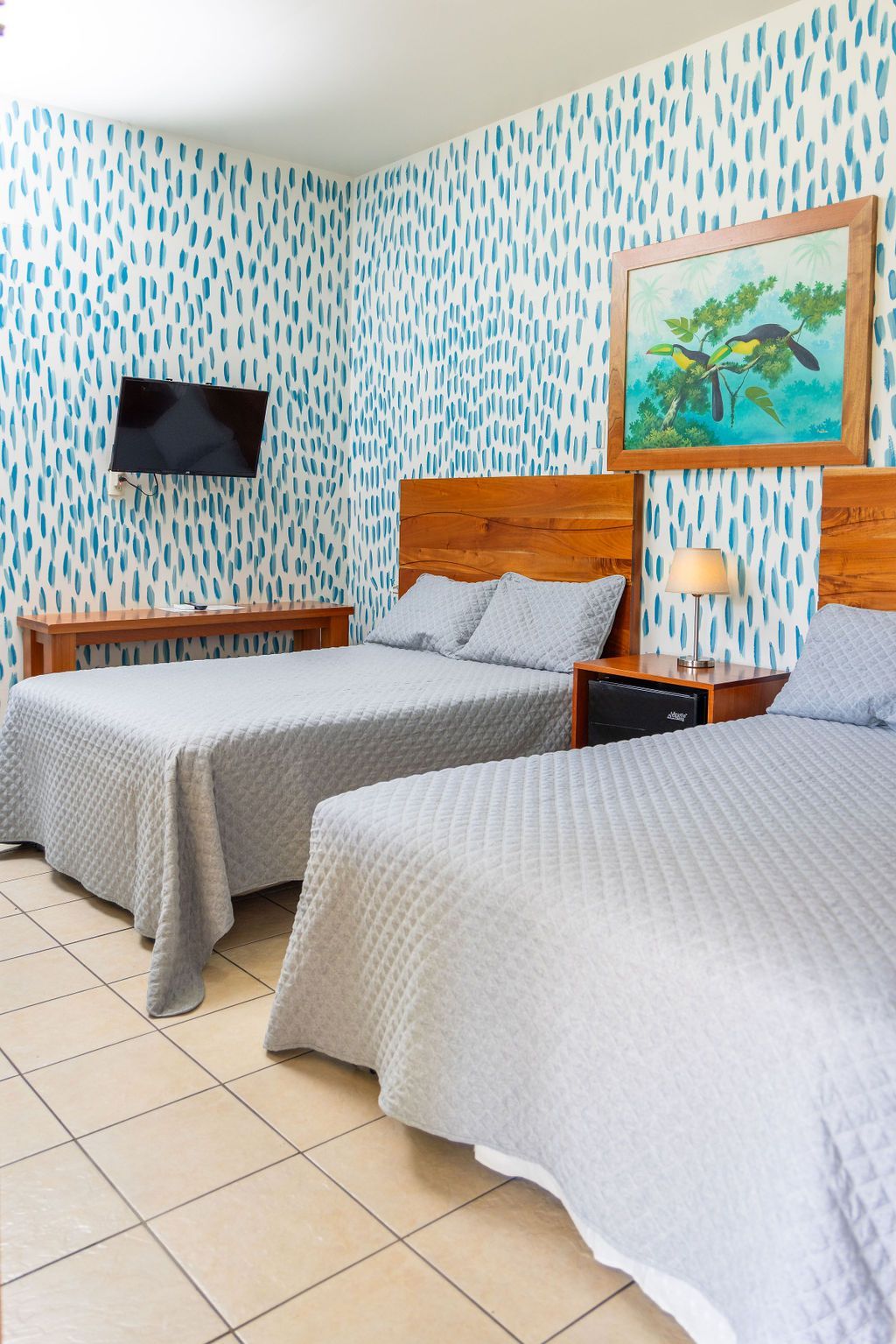 Dormitorio con dos camas, edredones grises, cabeceros de madera, papel tapiz estampado en azul, TV y obras de arte.