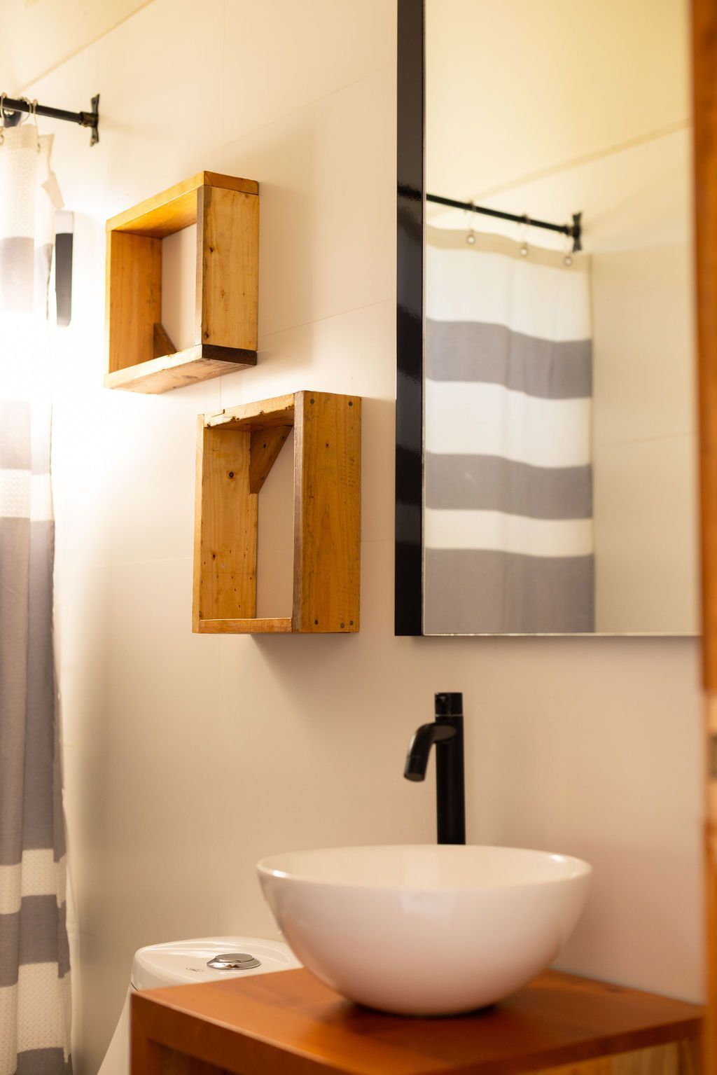 Baño con lavabo blanco, grifo negro, espejo, cortina de ducha a rayas y estantes de madera.