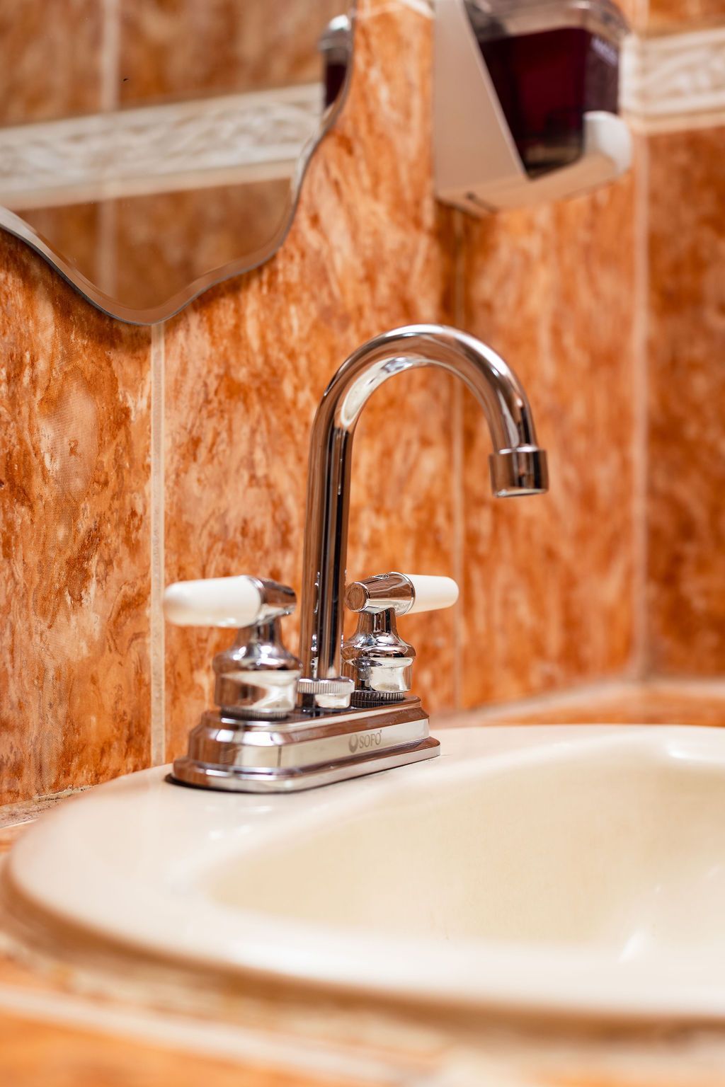 Grifo cromado con manijas blancas sobre un lavabo beige en un baño con azulejos naranjas.