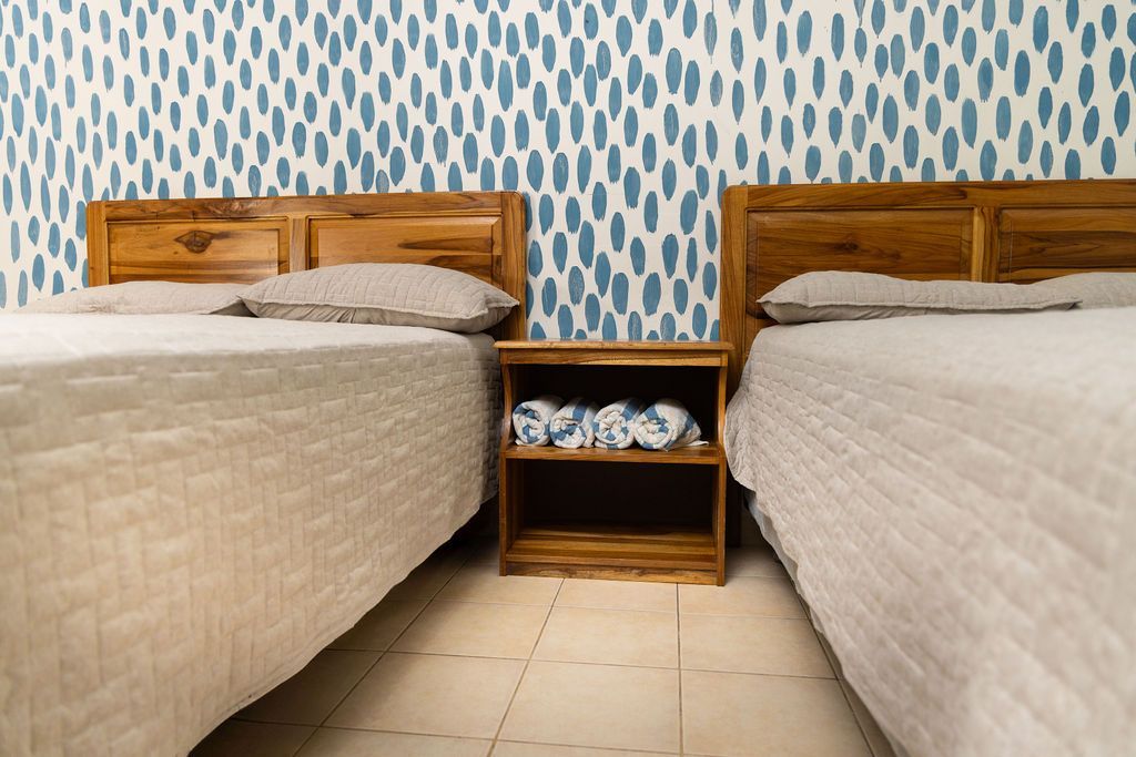 Dos camas de madera con ropa de cama blanca, mesita de noche pequeña, pared con estampado azul.