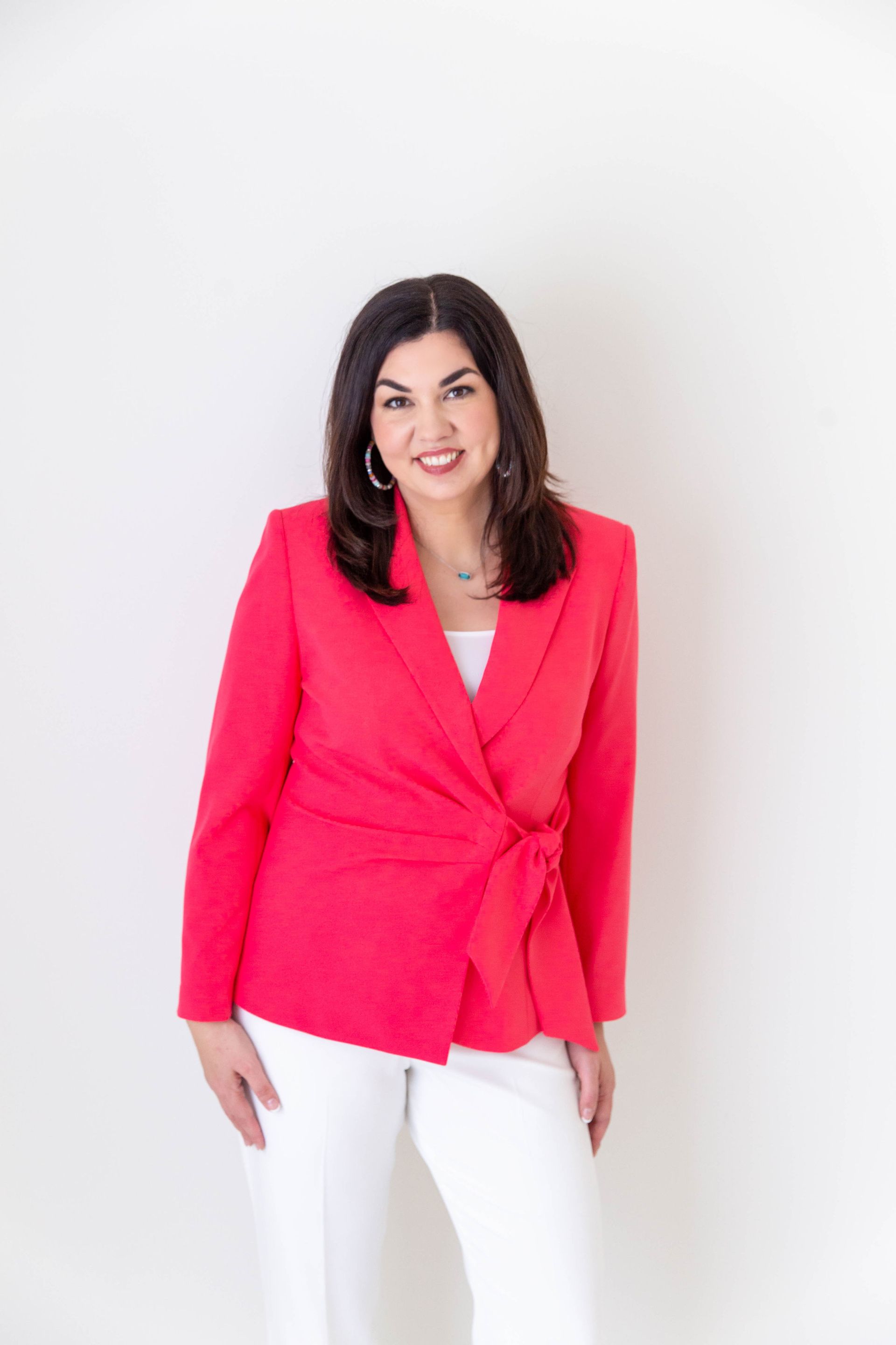 A woman in a red jacket and white pants is standing in front of a white wall.